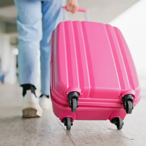 Getty pictures of a young woman wearing light jeans and white trainers withdraw a bright pink bag at the airport